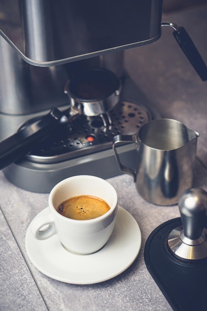 White cup with fresh hot coffee and milk on table near espresso\
machine pitcher and tamper