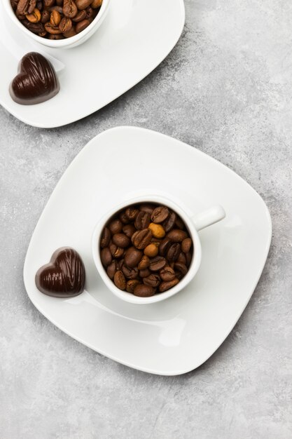 White cups for espresso filled with coffee beans and chocolate in form of heart on a light background. Top view.