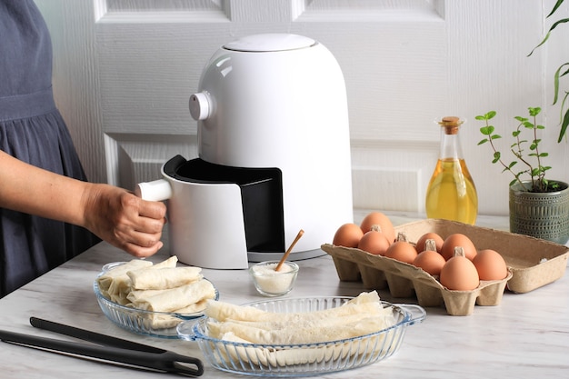 A white deep fryer or oil free fryer appliance, tongs, clear baking dish and egg tray are on the wooden table in the kitchen with a small plant in the pot (air fryer)