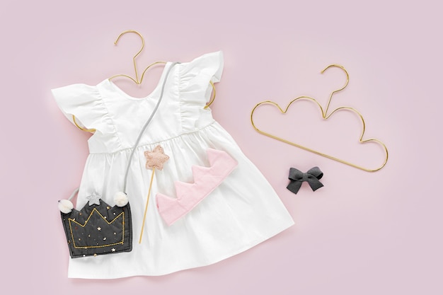 White dress with kids handbag shape of crown on golden hanger . Set of  baby clothes and accessories for spring or summer on pink background. Fashion childs outfit. Flat lay, top view
