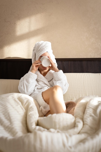 In a white dressing gown with a towel on her head a woman sits on the bed in the morning and drinks coffee