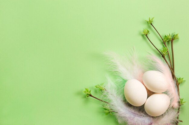 White easter eggs in a delicate nest of colored feathers with\
unfurled leaf buds