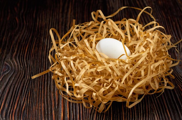 White eggs on paper straw in the shape of a nest on a wooden table. Low key.