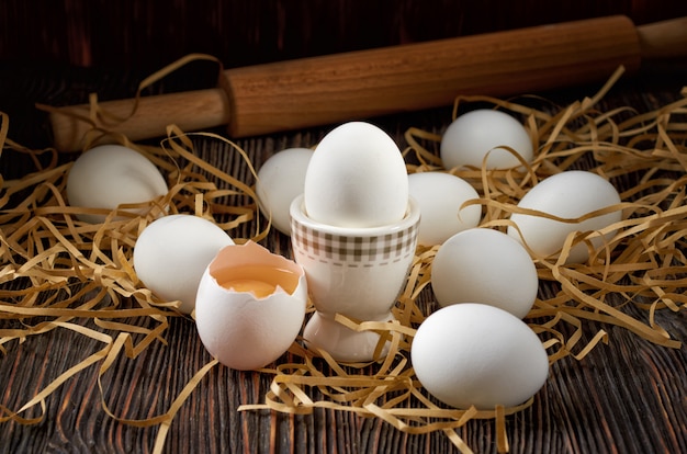 White eggs on paper straw and wooden table. Broken egg with yolk in the foreground. There's a rolling pin in the background. Low key.