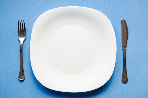 White empty dish on the blue tablecloth with fork and knife