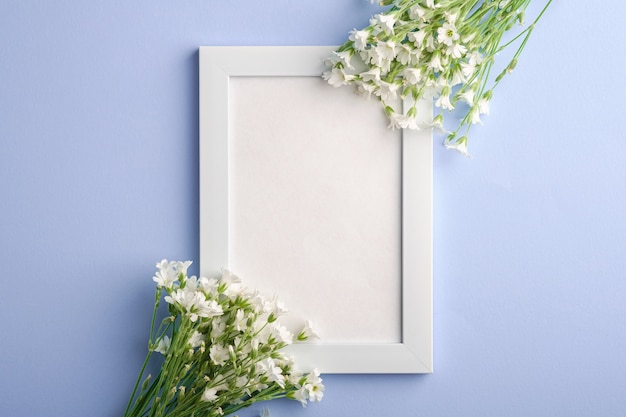 White empty photo frame mockup with mouse-ear chickweed flowers on blue background, top view copy space