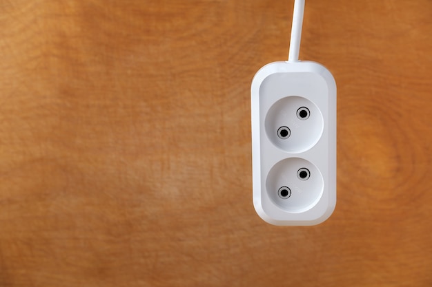 A white extension socket for two plugs hangs from top to bottom against the background of a textured and even wooden surface, and a copy space on the left.