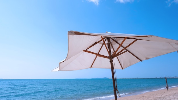 Ombrellone da spiaggia con trama in tessuto bianco e treppiede in legno marrone con nuvola bianca e cielo azzurro chiaro