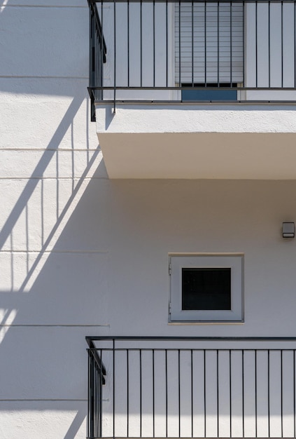 White facade of a flat building with minimalist design and
small windows the shadow of the black railing is projected on the
wall looking like stairs