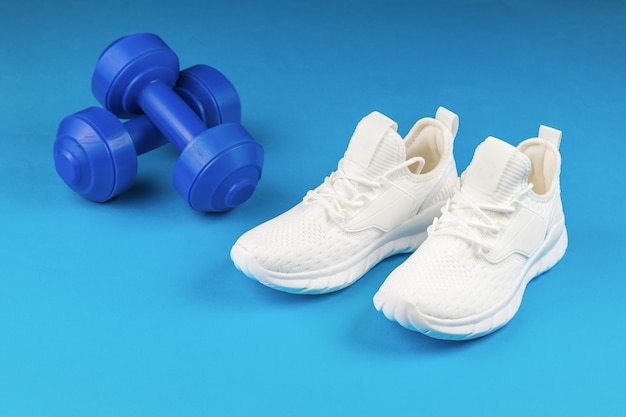 White fashionable fitness sneakers on a background of blue dumbbells on a blue background.