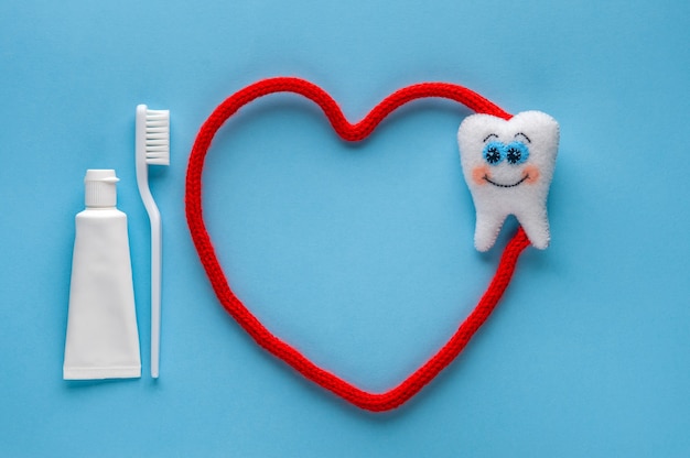 White felt tooth with toothpaste and toothbrush on blue background. Dental health concept with red heart. Flat lay, top view, copy space.