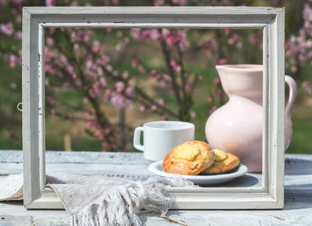 White frame, porcelain pink jug, cup and bakery on a table of white boards against the background of a flowering bush