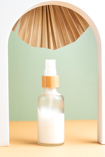 White frosted glass bottle of natural face moisturizer cream on beige background in white arch decorated with dry palm leaf Eco cosmetics concept