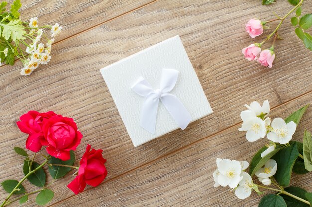 White gift box with beautiful roses, jasmine, peony and chamomile flowers on the wooden background. Concept of giving a gift on holidays. Top view.