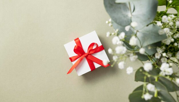 A white gift box with a red ribbon on a green background with a bouquet of eucalyptus and gypsophila. The concept of a holiday or gift card. Top  view and copy space.