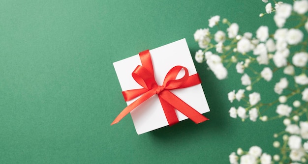 A white gift box with a red ribbon on a green background with a bouquet of  gypsophila. The concept of a holiday or gift card. Top  view and copy space.