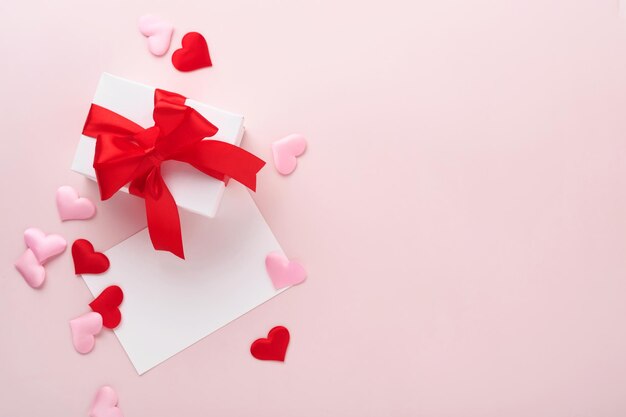 ピンクの背景に赤いリボンと小さな赤い装飾的なハートが付いた白いギフトボックス。コピースペースのある上面図。バレンタインデーや結婚式のロマンチックなコンセプト。お祭りの構成。モックアップ。