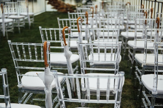 White glass chairs stand on the green grass round arch for a wedding ceremony in the background