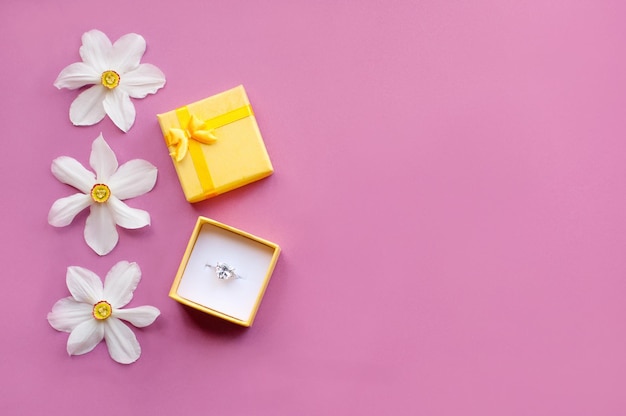 A white gold ring with a large gemstone in a yellow gift box on a pink background next to daffodil flowers with a place for text