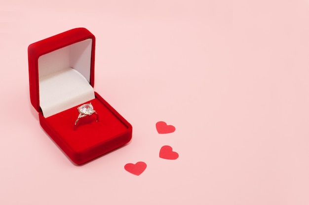 White gold ring with a precious stone in a red velvet box on a pink background with confetti in the form of hearts