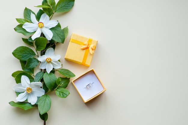 A white gold ring with a precious stone in a yellow open box with a bow lies on a beige background next to a branch with green leaves and white flowers with a yellow core with a place for text