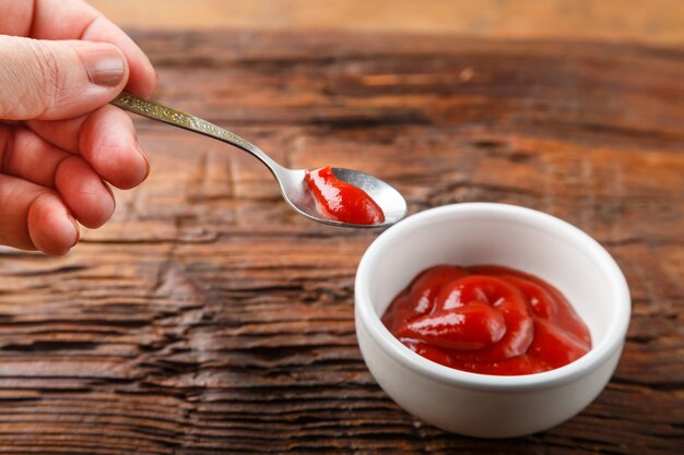 White gravy boat with ketchup and hand with a spoon of sauce on a wooden table. Horizontal photo.