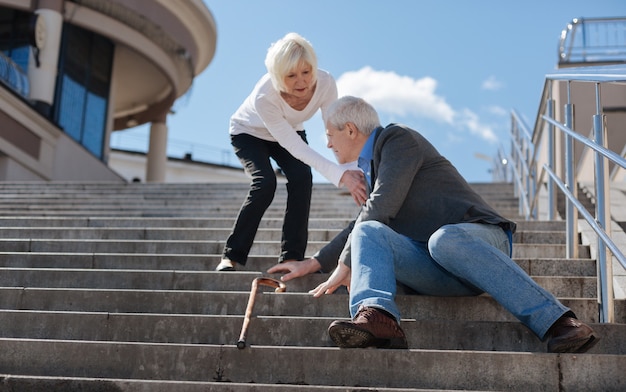 White-haired frustrated concerned pensioner sitting in the street and having dizzy while not indifferent woman helping him