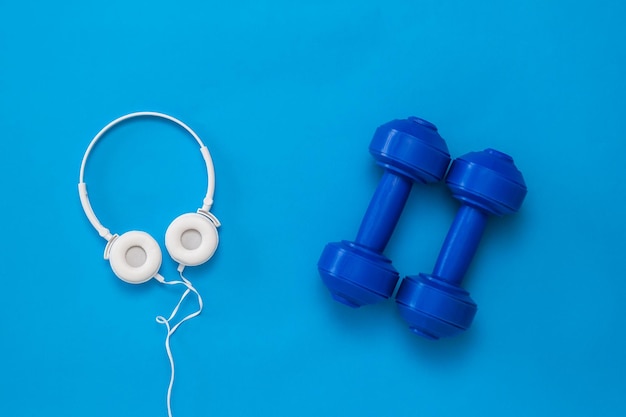 White headphones and blue dumbbells on a bright blue background. Accessories for fitness and weightlifting.