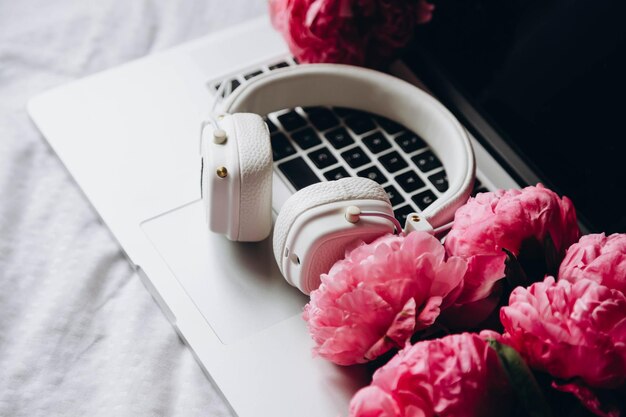 White headphones laptop and pink peonies flowers on white sheets in bed