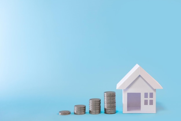 Модель белого дома со стопкой монет на синем фоне для управления денежными средствами, плана рефинансирования и концепции недвижимости.