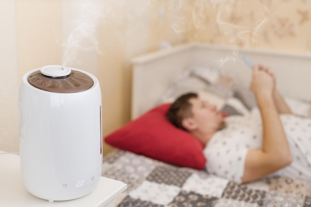 White humidifier spreading steam Humidification of the dry air in sleeping room Selective focus on vapor On the background young man chatting in smartphone Air purity and healthcare concept