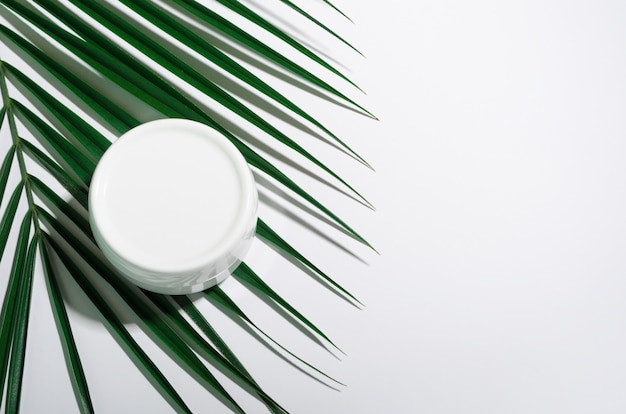 White jar of cream on a white table with tropical palm leaves.