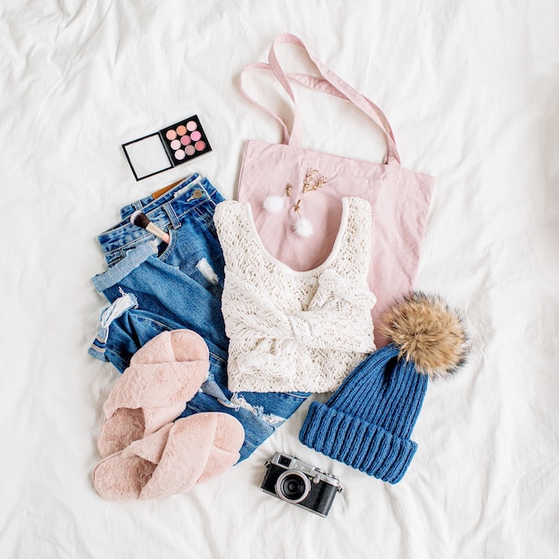 White knitted sweater, blue jeans, eco bag and hat on bed on white sheet. Women's stylish autumn or winter outfit. Trendy  clothes collage. Flat lay, top view.