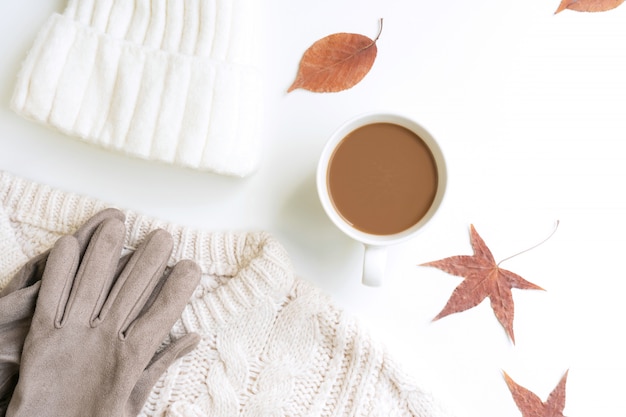 Maglione bianco lavorato a maglia, cappello di whool, una tazza di caffè, guanto e foglie di acero essiccate su un blackground bianco, piatto laico, vista dall'alto, copia spazio