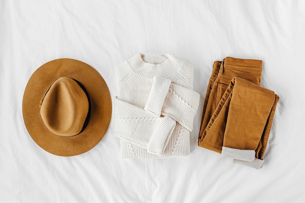 White knitted woolen sweater with brown trousers and hat on white bed. Women's stylish autumn or winter outfit. Trendy clothes collage. Flat lay, top view.
