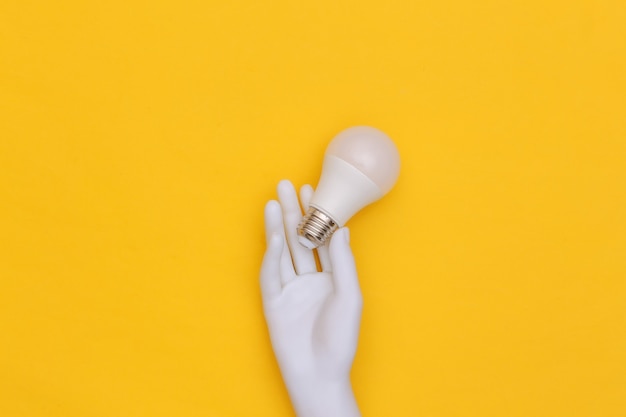 White mannequin hand holds LED light bulb on yellow background. Top view