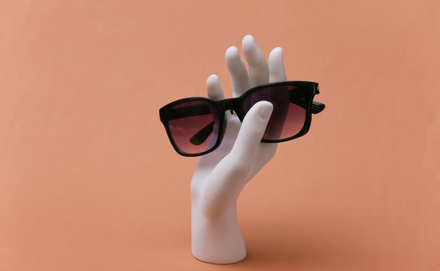 Белая рука манекена держит солнцезащитные очки на коричневом фоне