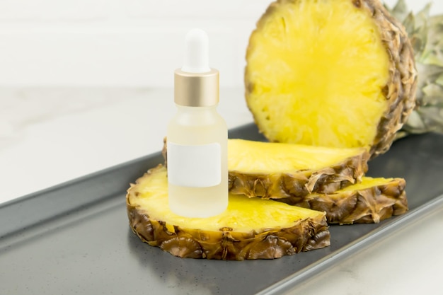 A white matte bottle stands on juicy pineapple slices and a ceramic dark tray. pineapple medicinal live cosmetics. Unbranded mock-up.