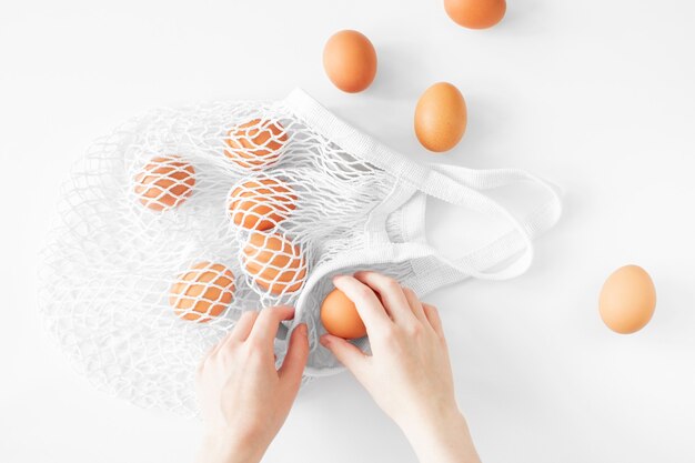 White mesh bag with brown eggs flat lay on white background top view copy space. Eco friendly, reusable shopping bag. Cotton knitted string bag. Zero waste easter concept. Minimalism style easter day