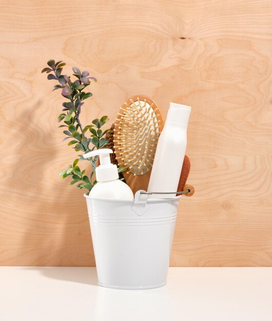 White metal bucket with wooden hairbrush liquid soap dispenser green branch on white surface