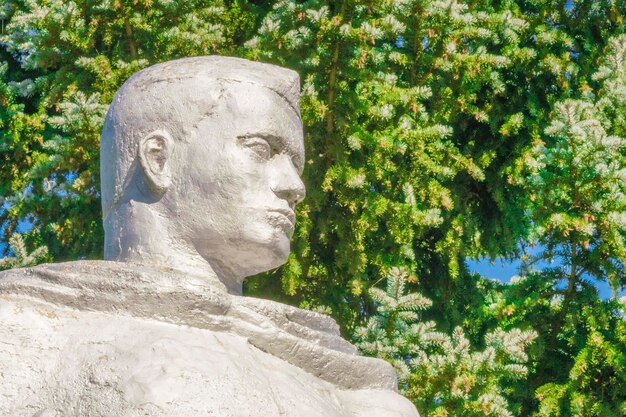 Белая металлическая статуя человека с оружием на фоне зеленых деревьев и голубого неба