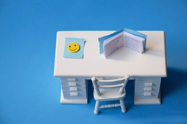 Foto scrivania da studio in miniatura bianca con un libro e una faccina sorridente motivante su sfondo blu