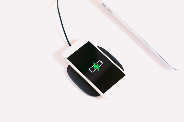 Smartphone mobile bianco con il logo di una batteria con un fulmine verde sullo schermo, in carica su una base di ricarica senza cavo accanto al laptop su un tavolo da lavoro bianco