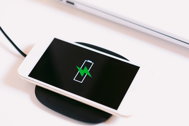 White mobile smart phone with the logo of a battery with green lightning on the screen, charging on a charger base without cable next to the laptop on a white work table