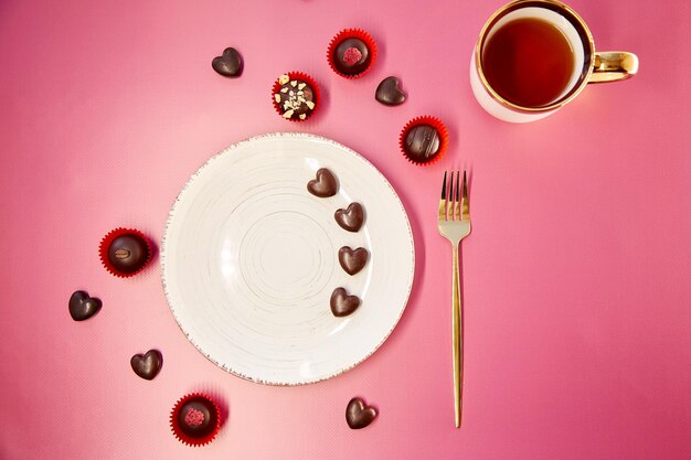 White mock up vintage plate with fork and cup of tea on pink gradient background Chocolate hearts and decorated sweets Romantic dinner Valentines Day concept View from above