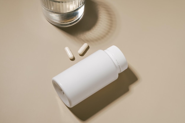 White mockup of a jar of pills and a glass of water on a beige background Concept of pharmaceutical products medical vitamins and dietary supplements