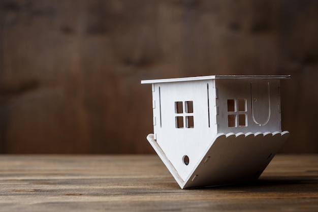 茶色の木製の背景に逆さまに三角形の屋根の上に立っている家の白いモデル。不動産のコンセプト
