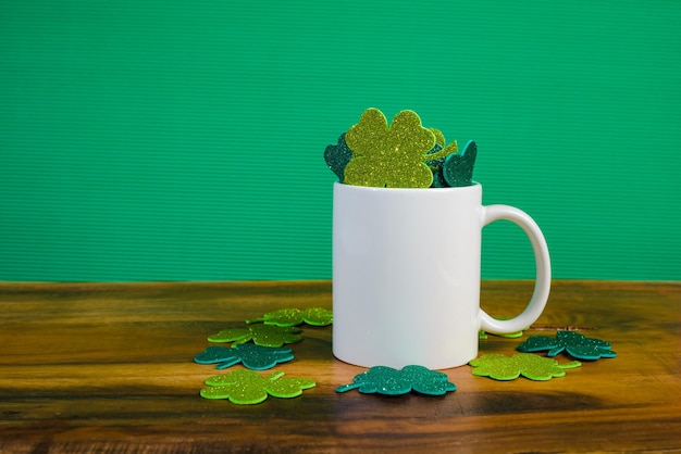 White mug full of clovers with glitter on wooden table Green background