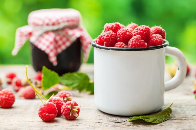 White mug of raspberries and jar of berry jam on background outdoorsxAxA