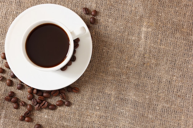 White mug with coffee, saucer and scattered coffee beans on rough burlap tablecloth, top view.